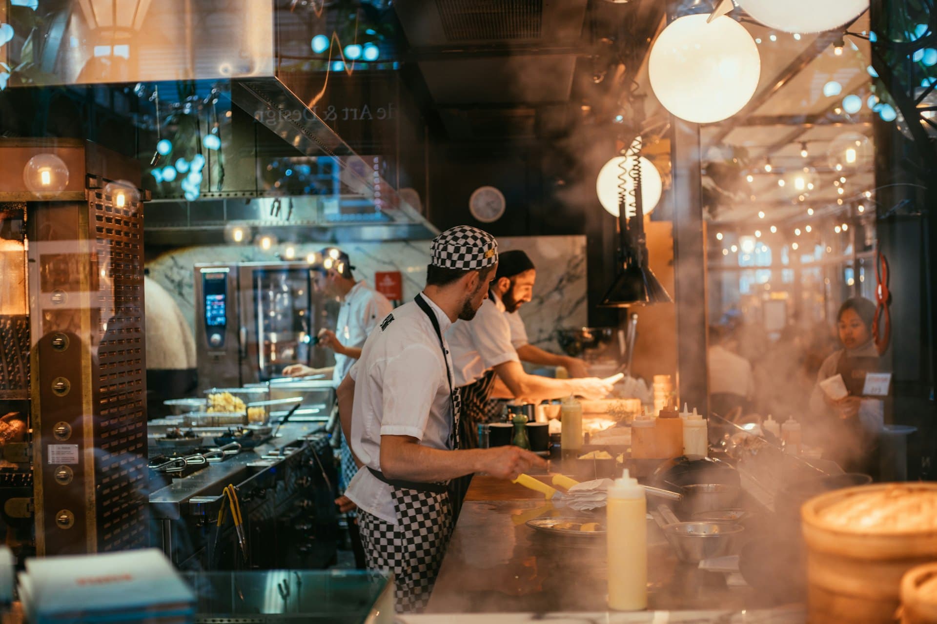 Cooks working a busy open kitchen pass under warm service lights, steam rising off the range